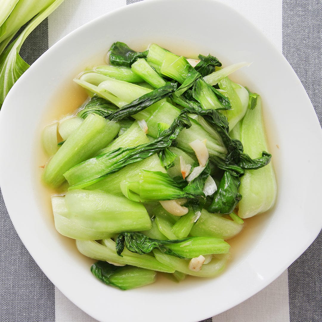 A bowl of cooked Asian greens made with 'White Stem' Organic Bok Choy from Back to the Roots in a light broth, garnished with sliced garlic, sits on a striped surface.