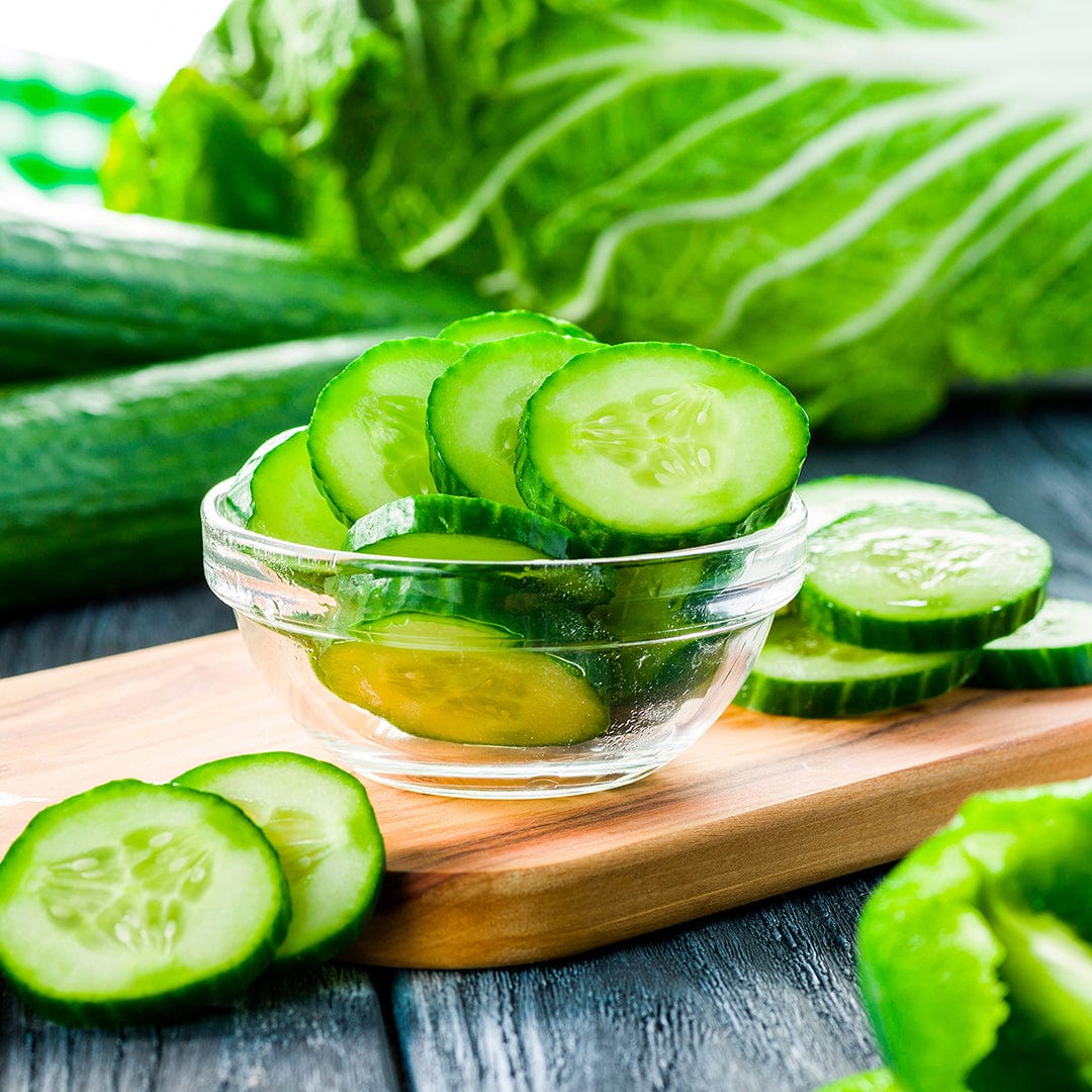 A glass bowl with sliced pickling cucumbers sits on a wooden board, whole cucumbers and greens nearby, grown from Back to the Roots Organic Cucumber Seeds—'Marketmore 76'.