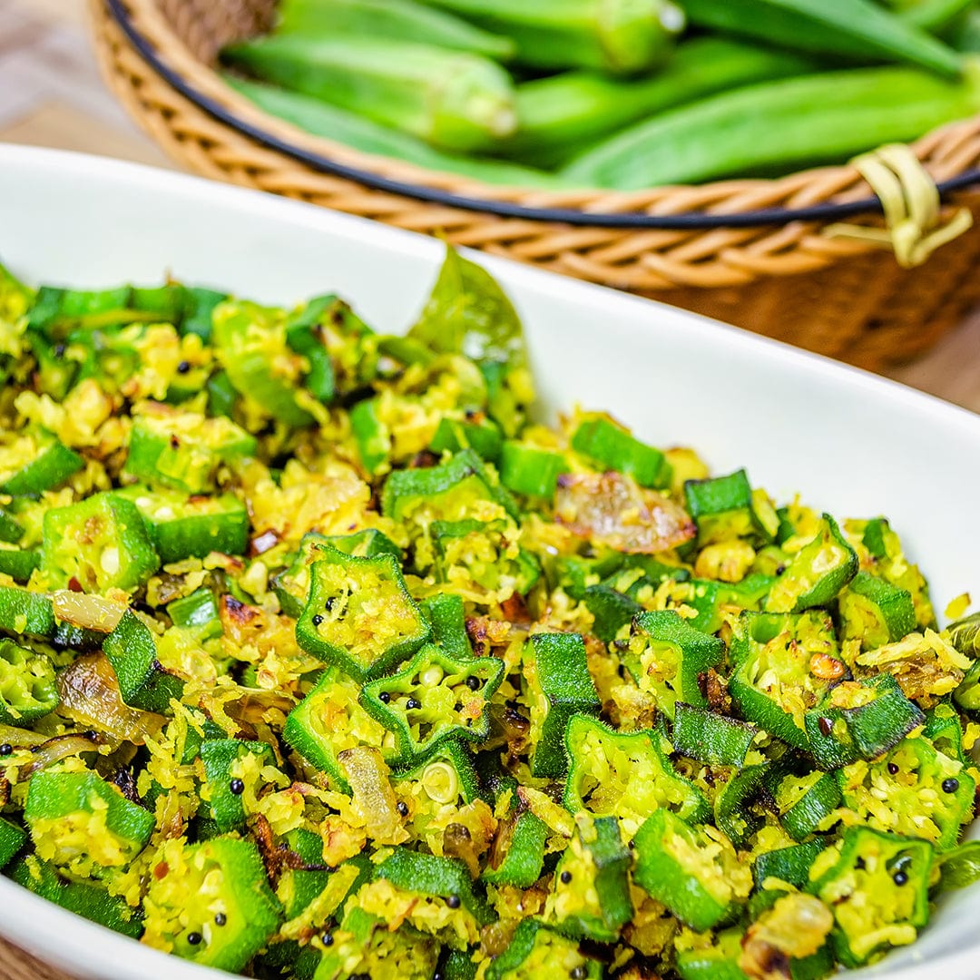 A white dish of spiced, cooked okra is shown with a basket of fresh pods grown from Back to the Roots Organic Okra Seeds (‘Clemson Spineless’) in the background.