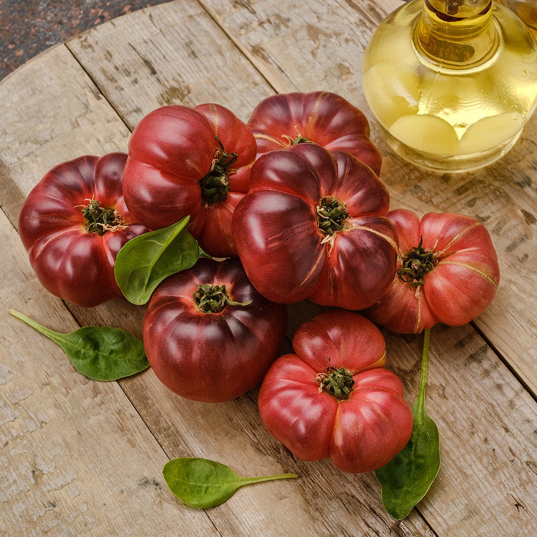 Cluster of large, ribbed Brandywine Pink tomatoes with deep red-pink hues on wooden surface with glass oil bottle. - Organic Tomato Seeds — 'Brandywine Pink' - Default Title - Back to the Roots