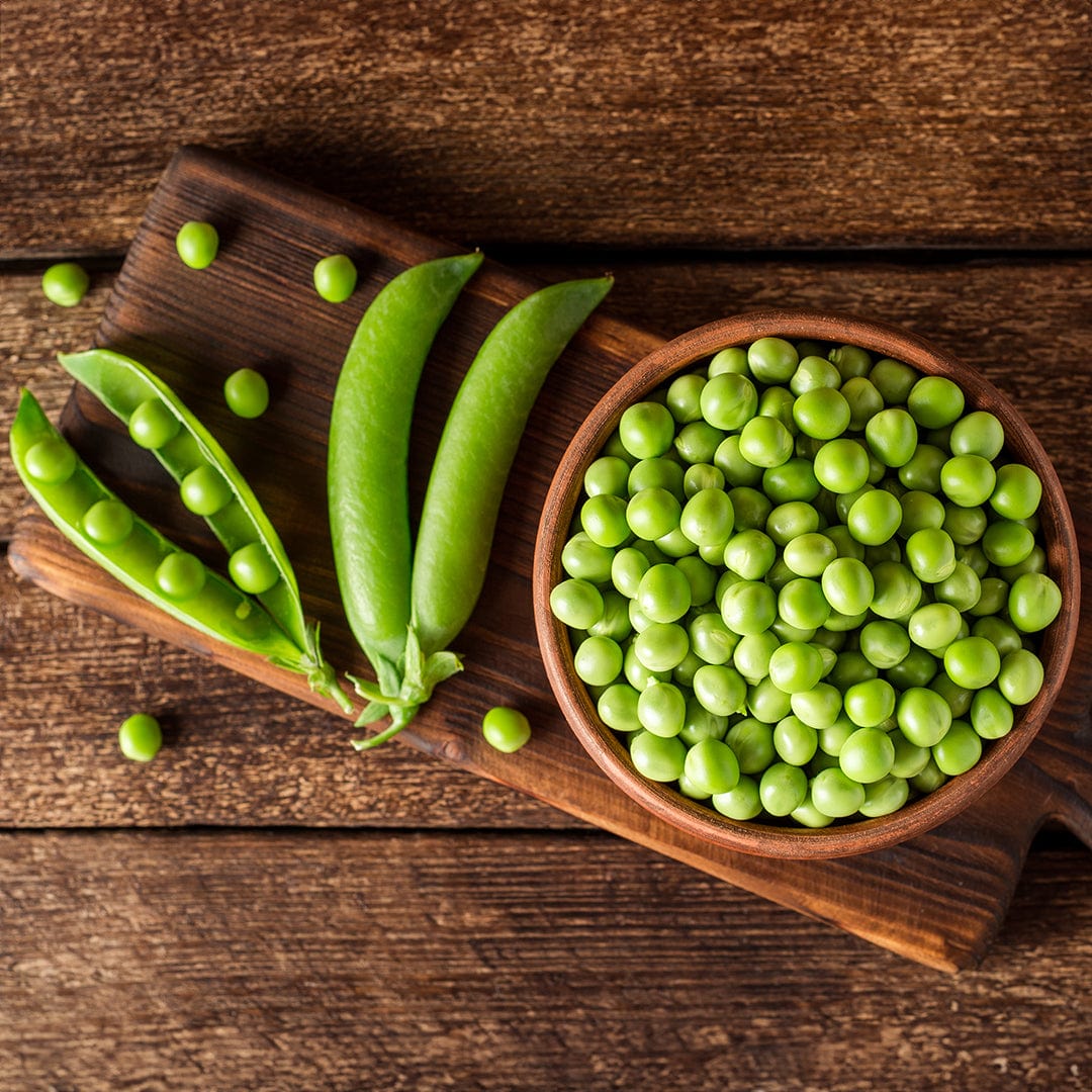 A wooden bowl of green peas—grown from Back to the Roots Organic Pea Seeds ‘Snap Sugar Daddy’—rests with pea pods and loose peas on a cutting board, ideal for any home gardener.