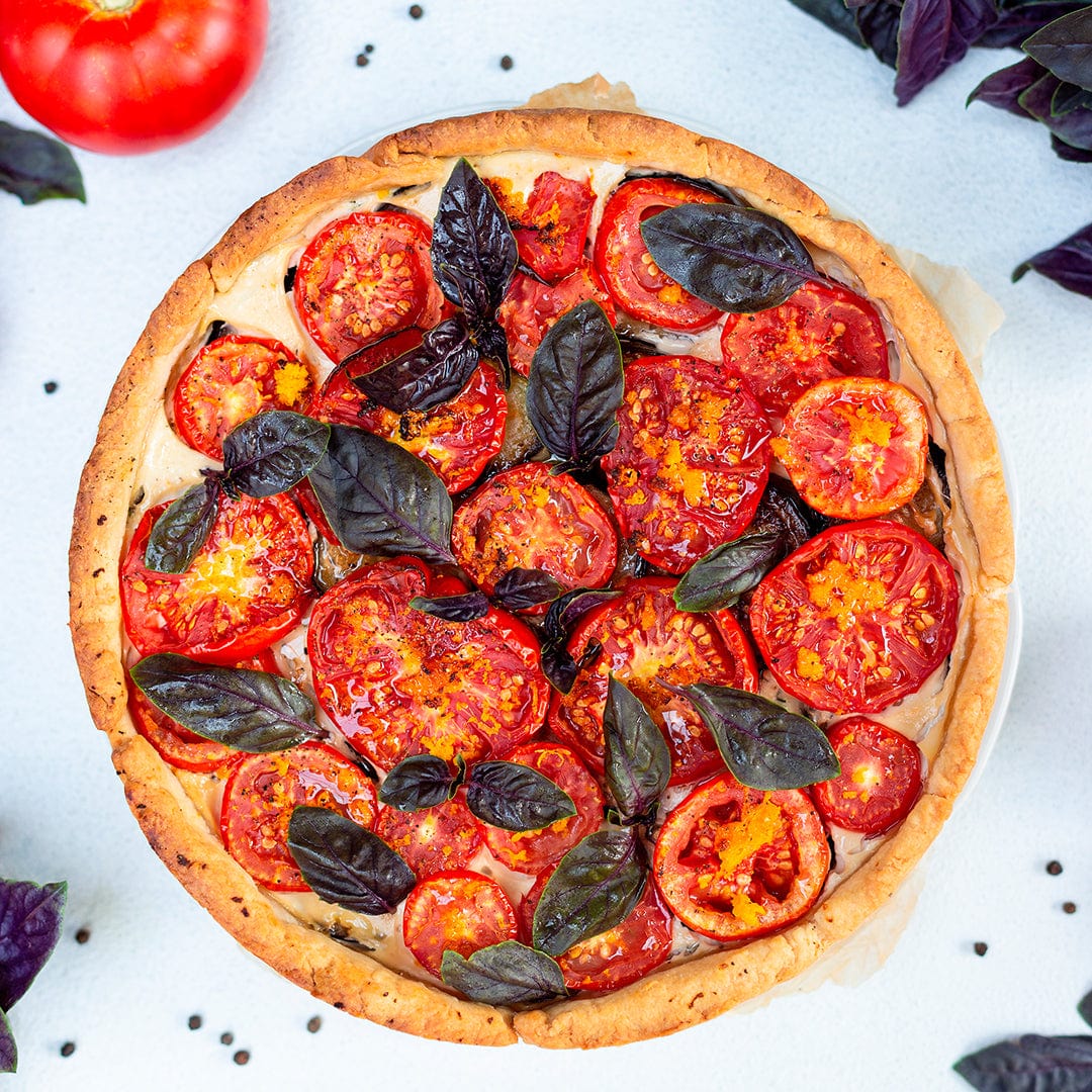 A tomato tart garnished with sliced tomatoes and Organic Basil Seeds — 'Red Rubin' by Back to the Roots, presented on a light surface with scattered peppercorns and basil.
