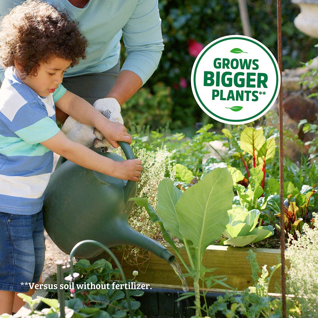 An adult and child water plants in a garden bed; overlay text: “Grows Bigger Plants*” and “Versus soil without Back to the Roots Natural & Organic ALL-PURPOSE Plant Food.”.