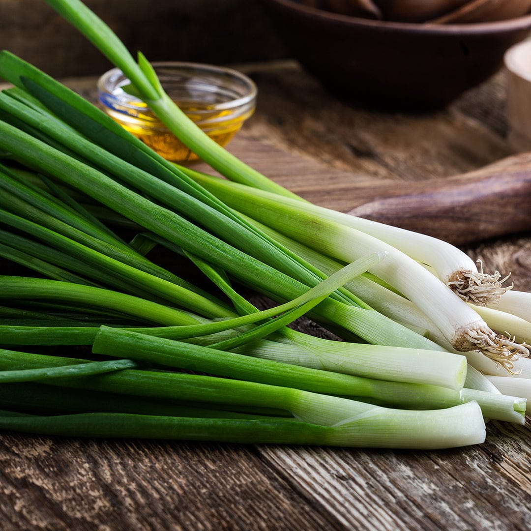 A bunch of green onions, grown from Back to the Roots' Organic Veggie Garden 5-Pack Seed Bundle, rests on a wooden surface with a bowl of oil in the background.