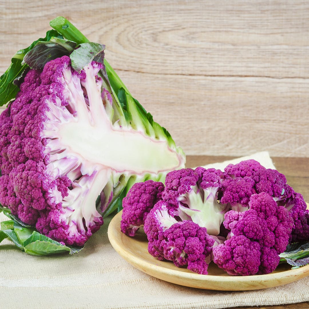 Vibrant purple cauliflower head with white stem, rough green leaves, and small florets arranged on a wooden plate. - Organic Cauliflower Seeds — 'Purple of Sicily' - Default Title - Back to the Roots