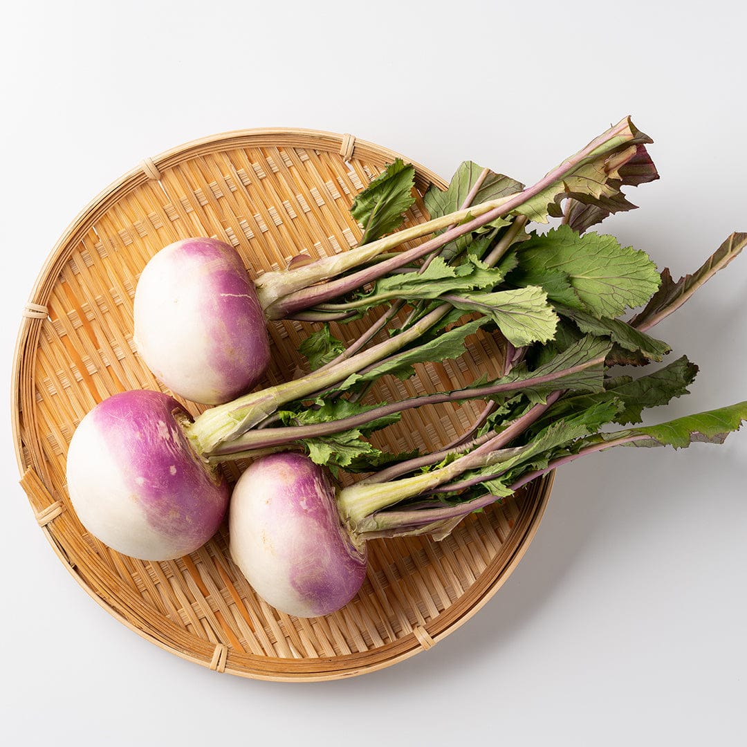 Three round turnips with purple tops and white bottoms, attached to leafy green stems, arranged on a woven bamboo plate. - Organic Turnip Seeds — 'Purple Top White Globe' - Default Title - Back to the Roots